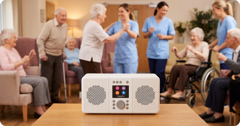 A group of residents and care staff dancing and laughing around a danceSing radio unit on a wooden table