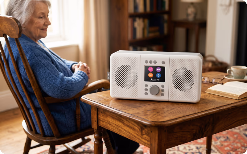 An elderly woman sitting calmly in a wooden chair beside a danceSing radio unit on a table